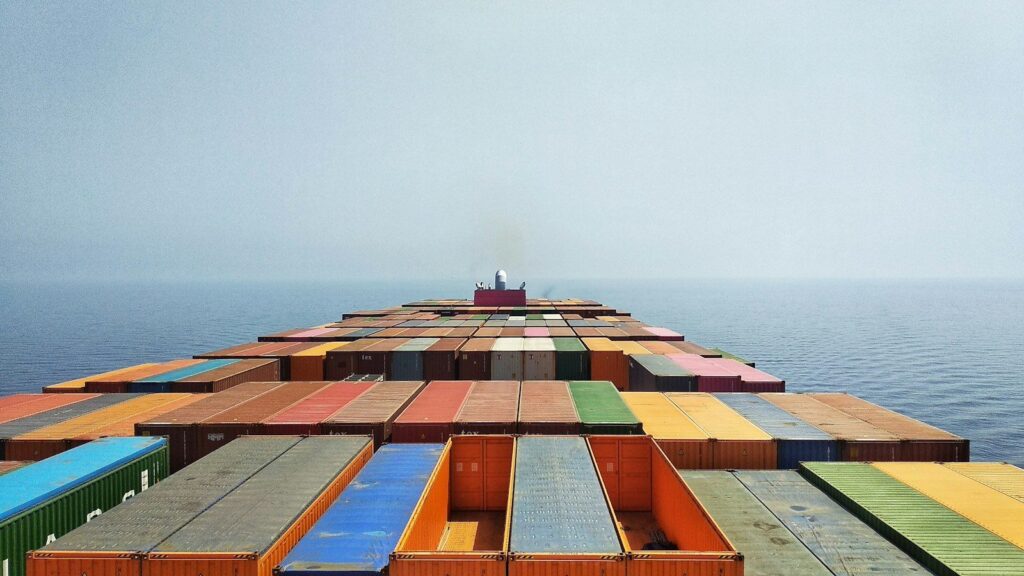 View across the container stacks of a loaded ship sailing on open sea toward a hazy horizon