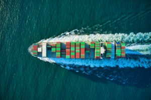Aerial view of a loaded container ship underway at sea, leaving a long white wake