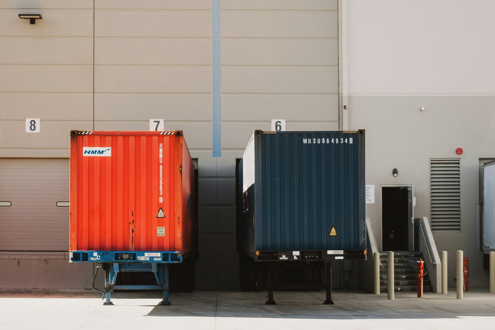 Two ocean containers on chassis backed into numbered warehouse dock doors at a distribution center